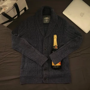 Mörkblå stickad kofta från H&M - Säljer en snygg mörkblå stickad kofta från H&M med knappar framtill. Koftan har ett diskret mönster och långa ärmar, perfekt för kyligare dagar. Passar bra till både vardag och mer uppklädda tillfällen. Nypris 1100