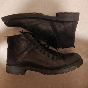 Svarta boots från Adidas - Snygga svarta boots från Adidas i skinn med snörning och dragkedja på sidan. Perfekta för en stilren look med robust sula för extra komfort. Passar bra till både jeans och mer uppklädda outfits.