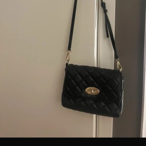 Svart quiltad axelväska - Snygg svart quiltad axelväska med gulddetaljer. Väskan har en praktisk dragkedja på baksidan och ett elegant spänne framtill. Perfekt för att bära dina nödvändigheter med stil.