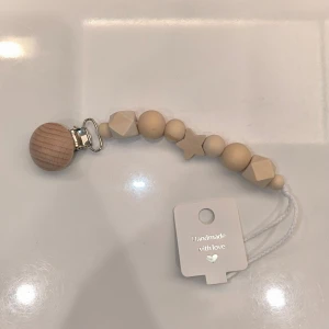 Napphållare med träpärlor - Säljer en handgjord napphållare med naturliga träpärlor i olika former, inklusive en stjärna. Den har en praktisk klämma i metall för enkel fastsättning. Perfekt för att hålla napp eller leksak nära till hands. Kan göra med namn vid beställning.