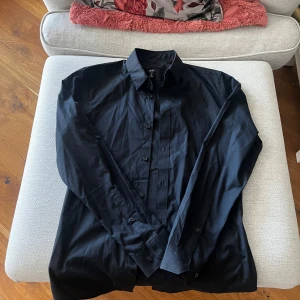 Svart skjorta från H&M - Snygg svart skjorta från H&M i slim fit. Perfekt för en stilren look med långa ärmar och knappar framtill. Materialet är lätt att stryka vilket gör den enkel att hålla snygg.