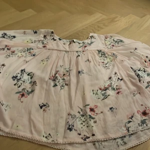 Ljusrosa blommig blus  från KappAhl - Säljer en söt ljusrosa blus från KappAhl med blommigt mönster. Blusen har en luftig passform och fina spetsdetaljer vid halsen. Perfekt för sommarens varma dagar!