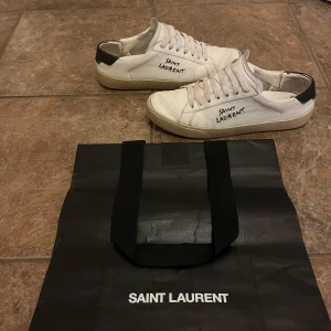 Vita sneakers från Saint Laurent - Snygga vita sneakers från Saint Laurent med svart detalj på hälen och handskriven logga på sidan. Skorna har en klassisk design med snörning och en robust sula. Perfekta för en stilren look. 41,5 passar upp till 42,5