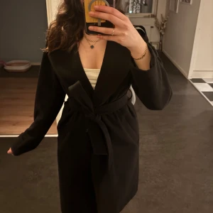 Svart kappa med knytband - Snygg svart kappa med knytband i midjan. Perfekt för att ge en stilren look. Den har långa ärmar och en klassisk design som passar till många tillfällen.