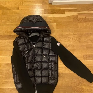 Moncler Cardigan  - Snygg svart quiltad jacka från Moncler med huva och dragkedja. Jackan har långa ärmar i ett annat material och en stilren design med röd och vit randdetalj på huvan. Perfekt för kyligare dagar.