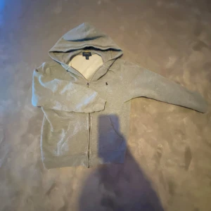 Grå hoodie från Ralph Lauren - Säljer en stilren grå hoodie från Ralph Lauren med dragkedja och huva. Den har ett diskret logotypbroderi på bröstet. Perfekt för en avslappnad look.