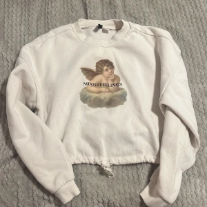 Beige croppad sweatshirt med ängelmotiv - Säljer en beige croppad sweatshirt med ett tryck av en ängel och texten 'Mixed Feelings'. Tröjan har långa ärmar och en justerbar dragsko i nederkanten. Perfekt för en avslappnad stil. Storlek M. Lite använd. 60% Bomull, 40% Polyester