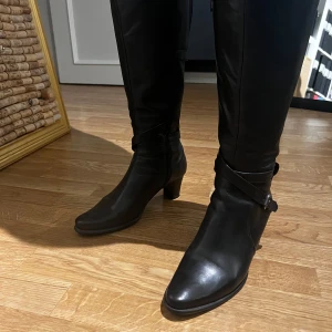 Svarta stövlar från Longo  - Snygga svarta stövlar i skinn med klack och dragkedja på sidan. De har en dekorativ rem med spänne runt vristen som ger en stilren look. Perfekta för både vardag och festliga tillfällen. Skick 9/10. Skriv vid intresse📩✨