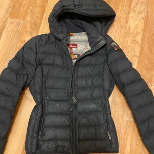 Svart dunjacka från Parajumpers - Säljer en svart dunjacka från Parajumpers med huva och dragkedja. Jackan har en patch på ärmen och är superlätt. Perfekt för kyliga dagar. Har även gått sönder längst ner ytterst lite och är ihopsytt men syns på nära håll
