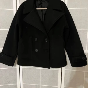 Design by si kappa med knappar - Snygg svart kappa med dubbelknäppning och bred krage. Perfekt för kyligare dagar med sin stilrena design och bekväma passform. Kommer tyvärr inte till användning🥰