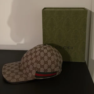 Beige keps från Gucci - Snygg beige keps från Gucci med det ikoniska GG-mönstret. Kepsen har en klassisk design med en justerbar rem baktill och en röd och grön randdetalj på sidan. Perfekt för att ge din outfit en lyxig touch.