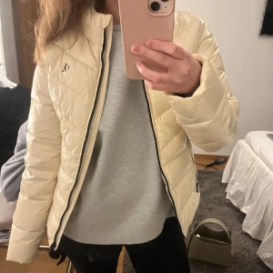 Beige-vit jacka från Calvin Klein - Säljer en snygg beige/vit jacka från Calvin Klein. Jackan har en dragkedja framtill och två fickor med dragkedjor. Ett glansigt material. Den har en logga på bröstet samt en svart detalj med märkesnamnet på ryggen. Nästan aldrig använd. 