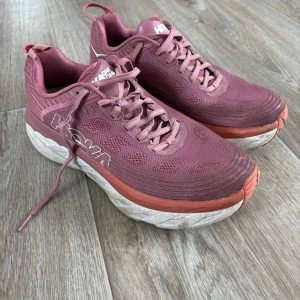 Rosa Hoka One One Bondi 6 - Säljer ett par rosa Hoka One One Bondi 6 sneakers med snörning. Skorna har en tjock sula och är designade för komfort och stöd. Perfekta för löpning eller promenader.