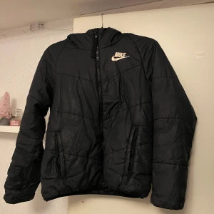 Svart dunjacka från Nike - Säljer en svart dunjacka från Nike med huva och dragkedja. Jackan har en stilren design med Nike-logga på bröstet. Perfekt för kyligare dagar.