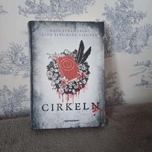Cirkeln - Boken 'Cirkeln' av Mats Strandberg och Sara Bergmark Elfgren är en spännande ungdomsroman. Den utspelar sig i Engelsfors där mystiska händelser inträffar. Omslaget är dramatiskt med en röd bok omgiven av blommor och fjädrar. Perfekt för dig som gillar fantasy och mystik.