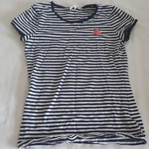 Randig t-shirt från Jonas Nielsen - Säljer en stilren randig t-shirt från Jonas Nielsen i blått och vitt. T-shirten har korta ärmar och en liten broderad detalj på bröstet. Perfekt för en avslappnad stil. Hör av dig vid intresse!