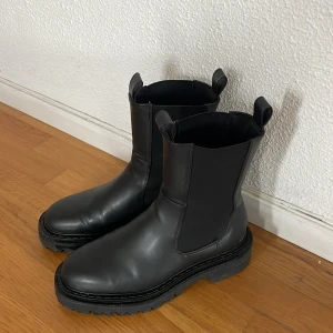 Svarta boots i vegan skinn - Snygga svarta boots i vegan skinn med robust sula och dragflikar upptill. Perfekta för en stilren look med en touch av edge. Står strl 37 men jag har 38