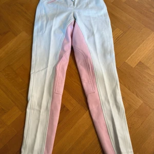 Vita och rosa ridbyxor - Snygga vita ridbyxor med rosa detaljer längs insidan av benen. Byxorna har en knapp och dragkedja framtill samt förstärkningar vid knäna för extra hållbarhet. Perfekta för tävling 