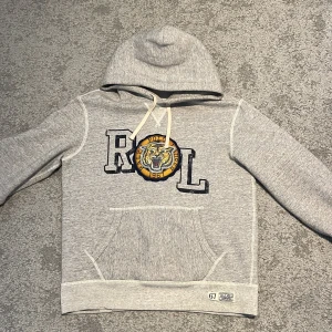 Grå hoodie från Polo Ralph Lauren - Säljer en snygg grå hoodie från Polo Ralph Lauren med en stor broderad tigerlogga och bokstäverna 'R L' på framsidan, del av designen att bokstäverna är lite slitna. 🤩                                                                                    Tröjan har en klassisk känguruficka och justerbar huva med snören. Perfekt för en avslappnad stil.                           Pris går alltid att diskutera! Vid frågor är det bara att höra av sig.😊