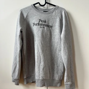 Grå sweatshirt från Peak Performance - Säljer en stilren grå sweatshirt från Peak Performance med tryck på bröstet. Tröjan har långa ärmar och ribbade muddar vid ärmslut och nederkant. Perfekt för en avslappnad stil.