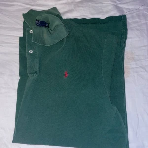 Grön pikétröja från Ralph Lauren - Säljer en klassisk grön pikétröja från Ralph Lauren med den ikoniska röda loggan broderad på bröstet. Tröjan har korta ärmar och knappar vid kragen. Perfekt för en stilren och avslappnad look. Den är väldigt stor skulle XL 