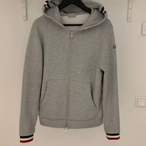 Grå hoodie från Moncler med dragkedja - Säljer en grå hoodie från Moncler med hel dragkedja och huva. Tröjan har två fickor framtill och Moncler-logga på ärmen. Ribbade muddar med röd, vit och marinblå rand vid ärmslut och nederkant. Perfekt för en avslappnad stil. Den har en liten fläck på armen som man knappt ser 