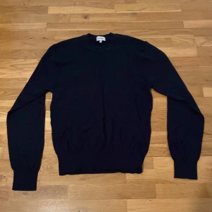 Mörkblå stickad tröja från Sandö - En klassisk mörkblå stickad tröja från Sandö i storlek S. Tröjan har rund halsringning, långa ärmar och ribbade muddar vid ärmslut och nederkant. Perfekt för lager-på-lager och enkel att matcha med olika outfits.