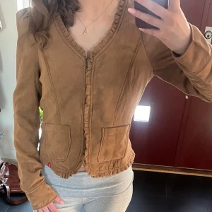 Brun mockajacka med volangkant - Säljer en brun mockajacka med volangdetaljer längs dragkedjan och nederkanten. Jackan har två fickor framtill och är figursydd med lång ärm. Perfekt för dig som vill ha en snygg och unik jacka till vardags.