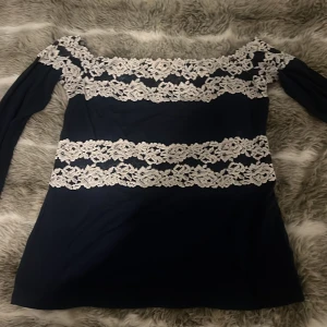 Marinblå långärmad topp med spets från Intimissimi Intimissimi - Mörkblå topp med långa ärmar och vita spetsdetaljer över bröst, midja och ärmslut. Snygg och feminin design med raka linjer av spets som ger en elegant touch. Perfekt för dig som gillar klassiska färger och fina detaljer. Och den säljs inte längreee