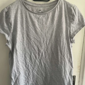 Ljusgrå t-shirt från H&M - En basic ljusgrå t-shirt från H&M i mjuk bomull. T-shirten har rund halsringning och korta ärmar, perfekt för en enkel och avslappnad stil. Passar dig som gillar klassiska och stilrena plagg.