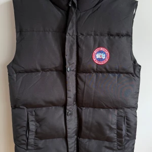 Svart dunväst från Canada Goose - Säljer en svart dunväst från Canada Goose med klassisk logotyp på bröstet och broderad märkning på ryggen. Västen har hög krage, två stora fickor framtill och stängs med dragkedja och knappar.