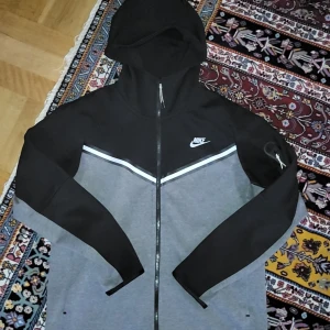 Svart och grå hoodie från Nike - Säljer en snygg svart och grå hoodie från Nike med dragkedja och huva. Jackan har en vit detalj över bröstet och Nike-logga på vänster sida. Perfekt för träning eller chill.