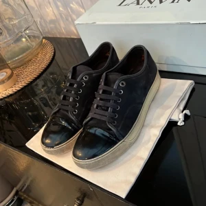 Svarta sneakers från Lanvin med lackad tå - Säljer ett par svarta sneakers från Lanvin med glans, Ny pris ungefär 4500kr. En bit av sulan har gått av där fram och det är därför jag säljer dom för 1200, man får med dustbag!