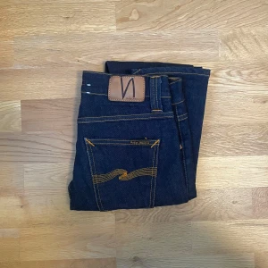 Mörkblå jeans från Nudie Jeans - Säljer ett par mörkblå jeans från Nudie Jeans med klassiska orange sömmar och bakfickor med vågig detalj. Modellen har normal passform