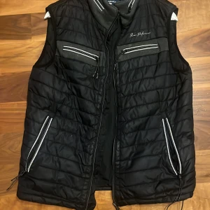 Svart quiltad väst från Performance - Säljer en svart quiltad väst från Performance med dragkedjor på fickor och bröstfickor. Västen har en sportig look med detaljer i grått och vitt samt ståkrage. Perfekt för lager-på-lager-stil.