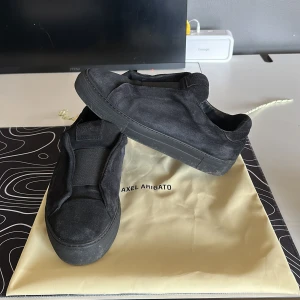 Svarta sneakers från Axel Arigato - Säljer ett par helsvarta sneakers från Axel Arigato med minimalistisk design. Skorna har platt sula, rund tå och är tillverkade i tygmaterial. Perfekta för en stilren och avslappnad look.