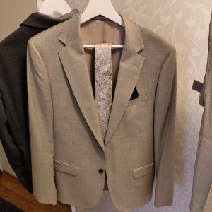 Beige kostym från Dressmann - Snygg beige kostym från Dressmann i 100% ull. Kavajen har klassisk passform med två knappar, fickor med lock och bröstficka. Byxorna har enkel design och matchar kavajen perfekt. Perfekt för bal eller student!