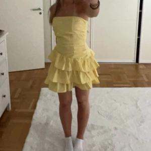 Gul volangklänning med axelbandslös design - Söt gul axelbandslös klänning med volanger i flera lager och kort längd. Klänningen har en figurnära passform upptill och utställd kjol. Storlek Xs