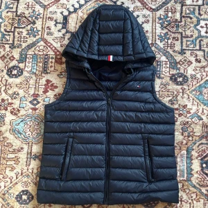 Marinblå dunväst från Tommy Hilfiger - Säljer en Marinblå dunväst från Tommy Hilfiger med avtagbar huva.                                                                  Med storleken M dom är små i storleken så passar XS till S