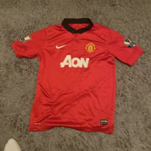 Manchester United hemmatröja 12/13 Van Persie - Manchester Uniteds officiella hemmatröja från säsongen 2012/2013 med Van Persie och nummer 20 på ryggen. Röd med svarta detaljer, Nike-logga och Aon som sponsor. Premier League-märke på ärmarna och autenticitetsmärke nertill. Som är köpt i england.