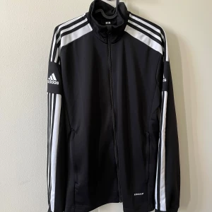 Svart Adidas träningsjacka med vita ränder - Klassisk svart träningsjacka från Adidas med vita ränder längs ärmarna och axlarna. Jackan har dragkedja framtill, ståkrage och ribbade muddar. Aldrig använd, skriv för mer info