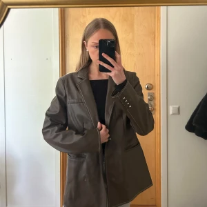 Brun skinnkavaj från NA-KD - Säljer en brun kavaj i skinnimitation från NA-KD. Kavajen har klassisk krage, långa ärmar och fickor framtill. Snygg passform och stilren design som passar till många outfits. Jättefint skick, utan skada eller förslitningar!