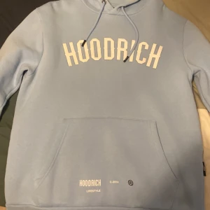 Ljusblå hoodie från Hoodrich - Säljer en ljusblå hoodie från Hoodrich med stor vit logga framtill och text på ryggen. Tröjan har huva med dragsko och en stor magficka. Perfekt för en avslappnad streetwear-look.