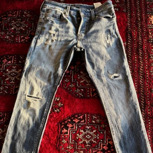 Ljusblå slitna jeans från Jack & Jones - Säljer ett par ljusblå jeans från Jack & Jones med slitningar och Klassisk femficksmodell med normal passform och dragkedja. Perfekta för en avslappnad stil. 