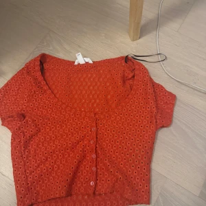 Röd kortärmad blus med broderade hål och knappar - Säljer en röd blus med korta ärmar och rund halsringning. Toppen har broderade hål över hela tyget och knäppning framtill. Perfekt för dig som gillar detaljer och färg!