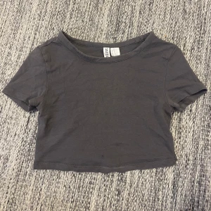 Mörkgrå croppad t-shirt - En enkel och mörkgrå croppad t-shirt från H&M. T-shirten har korta ärmar och rund halsringning. Den har två små fläckar på framsidan men det är inget man tänker på💗