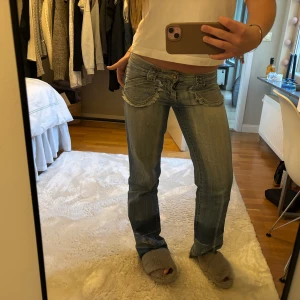 Lågmidjade bootcut jeans med strass och fransar - Säljer ett par ljusblå lågmidjade bootcut jeans från Big Bell. Jeansen har fransade fickor, strassdetaljer på både fickor och ben samt dekorativa sömmar. Perfekta för dig som gillar en unik och retro stil!