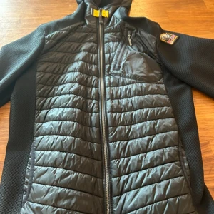 Svart hybridjacka Parajumpers - Säljer en svart hybridjacka från Parajumpers med quiltade partier framtill och baktill samt stickade ärmar och sidor. Jackan har dragkedja hela vägen, bröstficka med dragkedja och klassisk PJS-logga på axeln. Jackan är i nyskick.