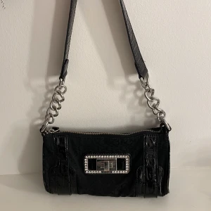 Svart axelväska från Guess med kedjedetaljer - Snygg svart axelväska från Guess med silverfärgad kedja och dekorativt spänne med strass framtill från 2007. Väskan har dragkedja upptill och detaljer i glansigt material och är äkta guess. Liten men praktisk. Knappt använd därav i bra kvalité. Går ej att få tag på längre.