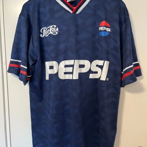Blå retro Pepsi jersey från Bershka - Snygg blå fotbollströja i retrostil från Bershka med stora Pepsi-loggor fram och bak samt nummer 24. Tröjan har korta ärmar och röd-vita detaljer på krage och ärmslut. Perfekt för dig som gillar vintage och sportig stil.